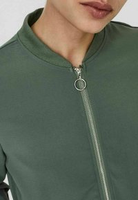Giacca bomber verde con collo a costine, dotata di una cerniera argentata e di un tessuto a trama liscia. Il cursore della cerniera ha un accento circolare.