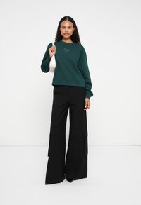 Donna che indossa un felpa verde scuro, pantaloni neri a gamba larga, scarpe nere e tiene una piccola borsa a tracolla bianca su uno sfondo bianco.