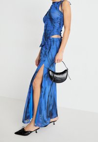 Niebieska sukienka maxi w wzory z bocznymi rozcięciami, cienkimi ramiączkami i marszczeniami, zestawiona z małą czarną skórzaną torebką i szpilkami.
