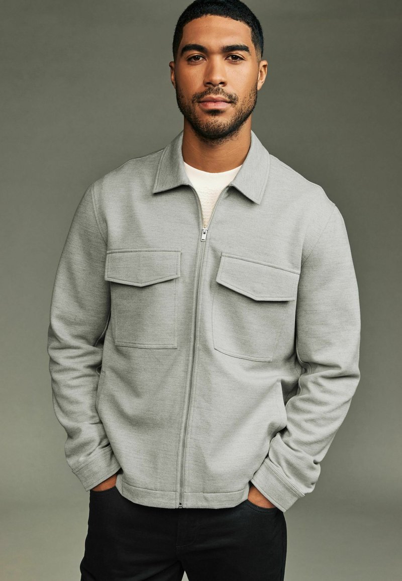 Light gray zip-up jacket with two front pockets, long sleeves, and a collared neck. Fabric appears soft and textured. Worn over a white shirt.