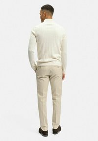 Krämfärgad kabelstickad tröja med hög krage, kombinerad med tanfärgade slim-fit byxor och bruna skor. Outfiten visar upp texturerat material och skräddarsydd design.