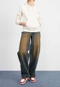 Sweat à capuche crème avec une poche avant, associé à un jean bleu ample avec un effet dégradé et des baskets marron avec des accents blancs.