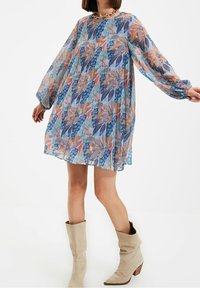 Chiffonkleid mit floralem Muster in Blau-, Pink- und Grüntönen. Lange Ärmel und eine lockere Passform, kombiniert mit beigen Knöchelstiefeln.