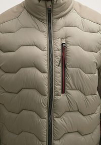 Lekka kurtka puchowa w stonowanym beżu, z pikowanym wzorem, wysokim kołnierzem, pełnym suwakiem z czerwonym akcentem i bocznymi panelami wentylacyjnymi.