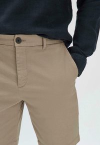 Beige shorts met een fitted ontwerp, riemlussen, voorzakken en een enkele knoopsluiting. De stof heeft een gladde, gestructureerde uitstraling.