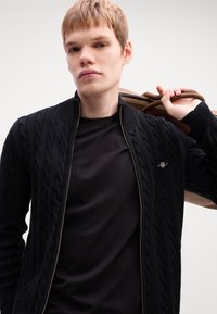 Fekete kötött kardigán, fonott mintával, cipzáros záródással és magas gallérral, fekete póló fölött viselve; a modell egy barna táskát hord.