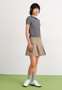 Csíkos fekete-fehér póló, khaki redőzött szoknya, szürke zokni és fekete-fehér tornacipő, síma fehér és zöld háttér előtt.