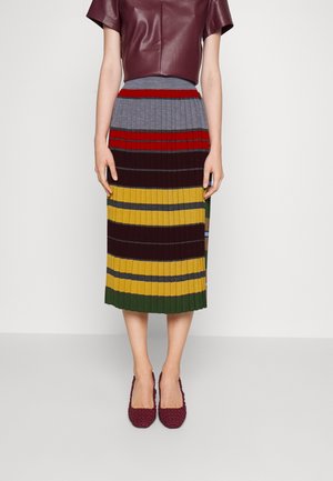 Femme portant une jupe plissée longueur genou avec des rayures horizontales en gris, rouge, marron, jaune et vert, associée à des chaussures bordeaux.