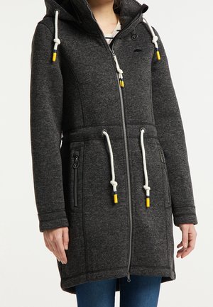 Manteau à capuche zippé gris foncé avec des cordons blancs ornés de pointes bleues et jaunes, porté sur un t-shirt rayé et un jean bleu.