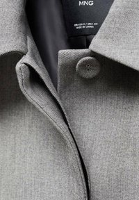 Camisa gris con cuello, hecha de tela texturizada, con cierre de botones y etiqueta en el interior. Vista en primer plano que resalta las costuras y el diseño del cuello.