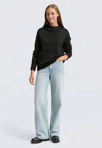 Juodas tekstūruotas megztinis su aukštu kaklu, derinamas su šviesiai mėlynais plačiomis kojomis džinsais ir juodais platforminiais batais. Modelis stovii ranka kišenėje.