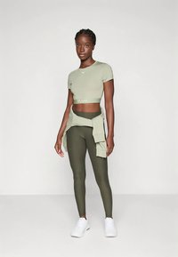 Top deportivo corto verde claro con logo, leggings oliva a juego y un suéter anudado. Zapatos deportivos blancos completan el conjunto.