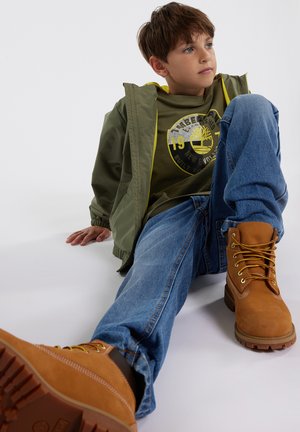 Jeune garçon assis par terre portant une veste verte, un t-shirt graphique vert, un jean bleu et des bottes à lacets beiges, regardant vers la droite.