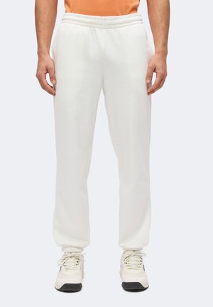 Man draagt witte joggerbroek met elastische taille en witte sneakers, staat met ontspannen handen langs het lichaam.