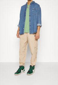 Denimskjorta med knappstängning över en grön T-shirt, beige texturerade cargobyxor och högsockade gröna och svarta sneakers med vita detaljer.