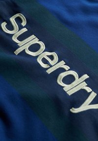 Niebieska i ciemnozielona tkanina z haftowanym logo "Superdry" w jasnej kremowej barwie. Charakteryzuje się gładką fakturą oraz poziomymi paskami kolorów.