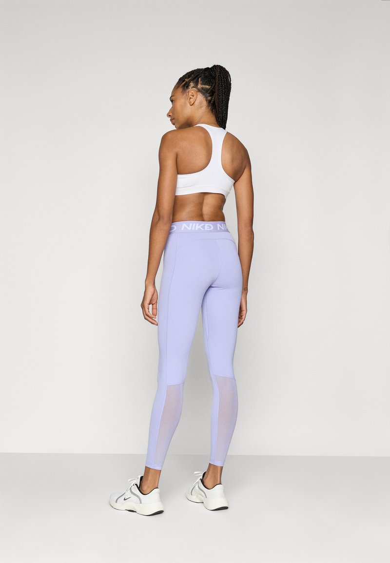 Leggings de color púrpura claro con paneles de malla y una cintura alta que presentan un logo de Nike en blanco, combinados con una camiseta deportiva blanca y zapatillas.