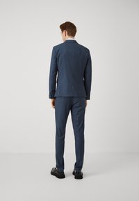 Sötétkék kockás öltöny, testre szabott fazonnal, dupla hasítékos zakóval és slim-fit nadrággal, fekete cipővel párosítva.