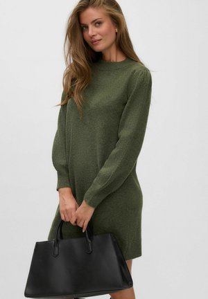 Femme aux longs cheveux portant une robe verte à manches longues, tenant un grand sac à main en cuir noir sur un fond clair uni.