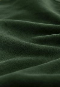 Mörkgrön tyg med en mjuk, matt textur, som visar släta, svagt vågiga veck och subtila variationer i ton och djup.