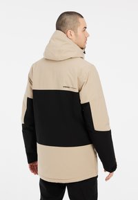 Beige en zwart gekleurde blokjasje met een capuchon, ritsopening en verstelbare zoom. Gemaakt van weerbestendig materiaal met logodetail.