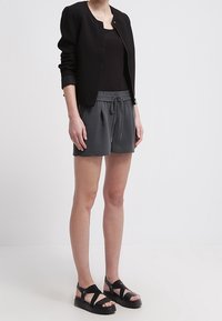 Zwarte cropped jas over een mouwloos donker tanktop, gecombineerd met grijze short met trekkoord en zwarte sandalen met bandjes. Gladde stof, ontspannen pasvorm.