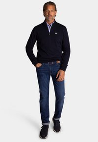 Marineblauwe gebreide trui met een halve rits en kraag, gecombineerd met blauwe denim jeans en donkere sneakers, met een gestructureerde stof en casual ontwerp.