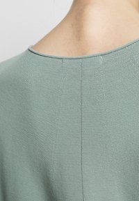 Gros plan sur le haut du dos et le cou d’une personne portant une chemise tricotée texturée vert clair avec un col rond.