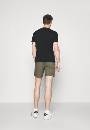 Homme de dos portant un t-shirt noir, un short vert olive, des chaussettes blanches et des baskets blanches avec des accents verts foncés, debout devant un fond uni.