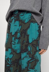 Grijze gebreide trui die gedeeltelijk een groene en turquoise rok met grote bloemmotieven en contrasterende zwarte contouren bedekt.