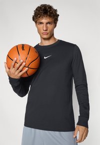 Langærmet sort Nike skjorte med hvid logo, parret med grå shorts. Modellen holder en orange basketball med sorte detaljer.