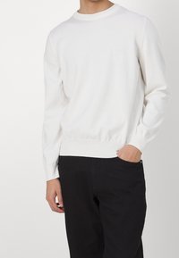 Weißer Rundhalsausschnitt-Pullover aus weichem Material mit gerippten Bündchen und Saum. Trägt man mit schwarzen Hosen, subtil sichtbares Markenlogo auf der Brust.
