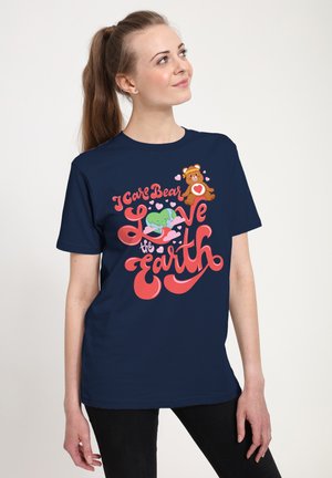 Kvinde iført mørkeblå t-shirt med farverig tekst "I Care Bear Love the Earth", hjerte, bamse og jordklode illustrationer.