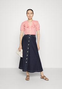 Blusa rosa clara com decote em babados, combinada com uma saia maxi de denim escura com fecho frontal por botões e bolsos laterais. Sandálias deslizantes marrons.