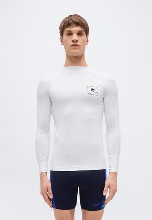 Νεαρός άνδρας που φοράει λευκό μακρυμάνικο rash guard με λογότυπο και μαύρο μαγιό με μπλε ρίγες στο πλάι και μικρό λογότυπο μάρκας.