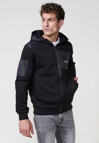 Sudadera con cremallera negra, textura acanalada, capucha y paneles de tela variados. Cuenta con bolsillos con cremallera en el pecho y los brazos, puños elásticos y dobladillo.