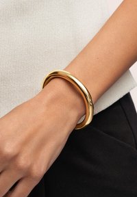 Pulsera de bangle de metal dorado con un diseño suave y redondeado, descansando sobre una muñeca. La superficie brilla, reflejando la luz.