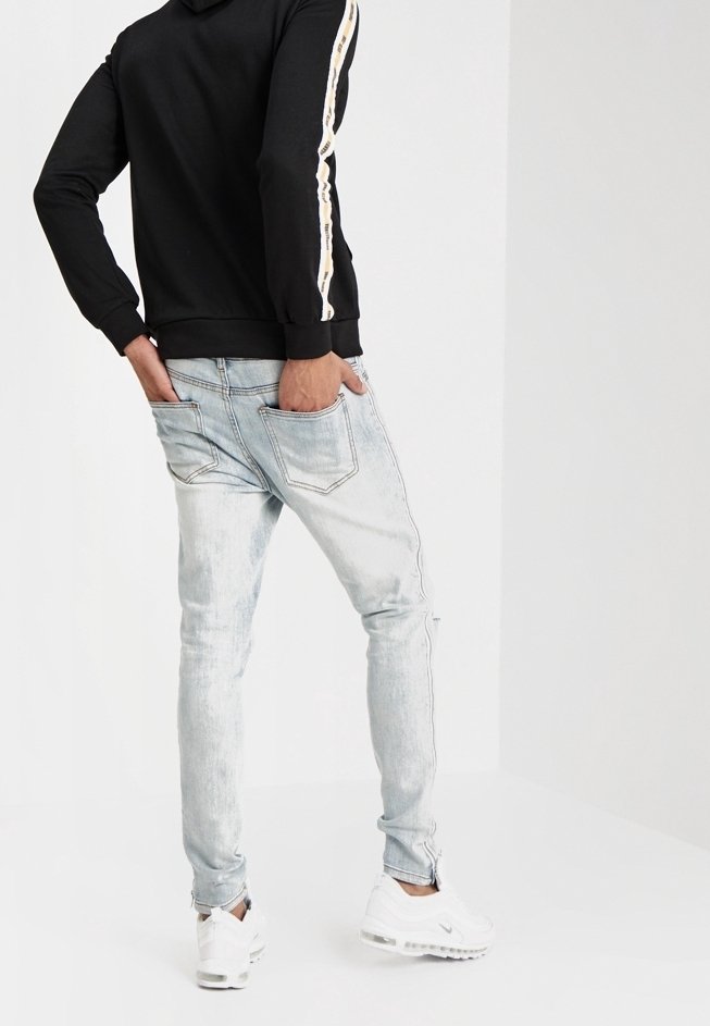 Schwarzer Kapuzenpullover mit weißen und goldenen Seitenstreifen, kombiniert mit hellblauen engen Jeans und weißen Sportschuhen. Lässiger Streetwear-Stil.
