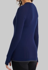 Tmavě modrý top s dlouhým rukávem, přiléhavým střihem, texturovanou látkou a jemnými vzory. Má otvory na palce na manžetách a zakřivený lem.