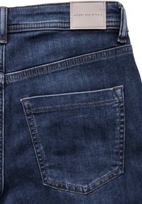 Jeans de mezclilla con un lavado azul oscuro, que presentan una etiqueta de cuero con "Street One Studio" y un solo bolsillo trasero con costuras visibles.