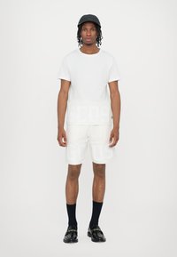 T-shirt in cotone bianco con un audace pattern del logo GCDS. Abbinato a pantaloni corti bianchi con un marchio simile. Un berretto nero e sofisticate scarpe loafers completano il look.