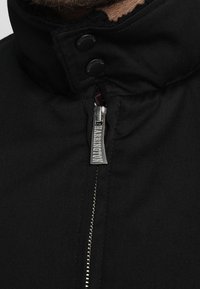Black jacket featuring a high collar, silver zipper with a tag, and two snap buttons at the collar. Durable fabric with a smooth texture.