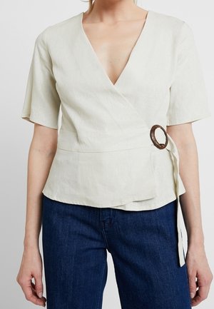 Vrouw draagt een crèmekleurige korte mouw overslagblouse met een ronde houten gesp en donkerblauwe spijkerbroek.