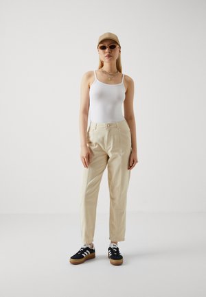 Pantalones beige de cintura alta con un corte relajado, combinados con una camiseta sin mangas blanca. Los accesorios incluyen una gorra beige y zapatillas negras con rayas blancas.
