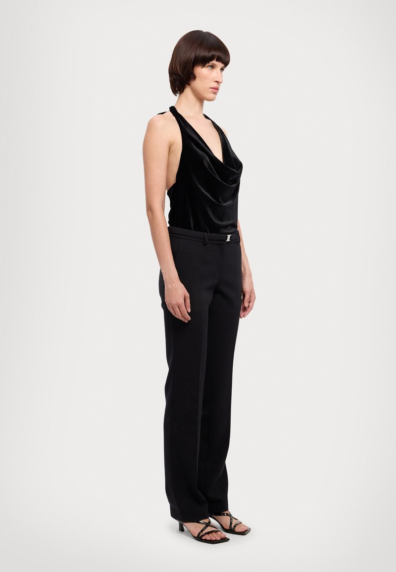 Top de veludo preto com gola cowl e alças halter, combinado com calças pretas ajustadas com um cinto metálico. Sandálias pretas de tiras.