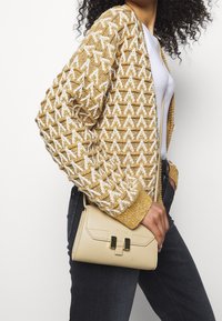 Borsa in ecopelle beige di forma rettangolare, con dettagli in metallo dorato e chiusura a scatto, tenuta accanto a un cardigan texturizzato giallo senape e bianco.