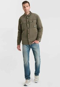 Man in een olijfgroene corduroy jas, lichte spijkerbroek en witte sneakers, staand tegen een witte achtergrond.
