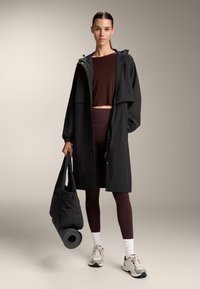 Nainen, joka on pukeutunut mustaan sadetakkiin ruskeiden legginsien ja crop-topin päälle, pitää mustaa joogamaton laukku kädessään ja seisoo neutraalilla taustalla.