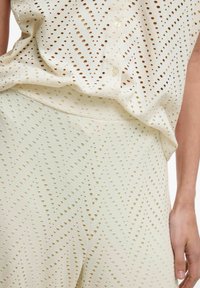 Gaiši bēšs komplekts, kas ietver topu un bikses ar perforētu mazu apļiņu un chevron tekstūru, veicinot gaisa caurlaidību un dizaina estētiku.