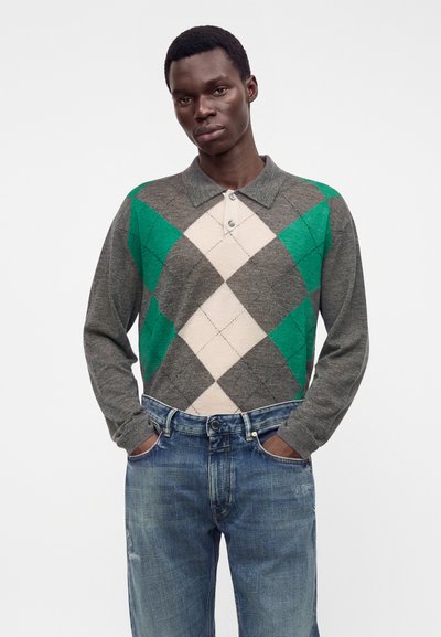 Homme portant un pull polo à manches longues argyle gris, vert et beige, avec un jean bleu, debout les mains dans les poches.